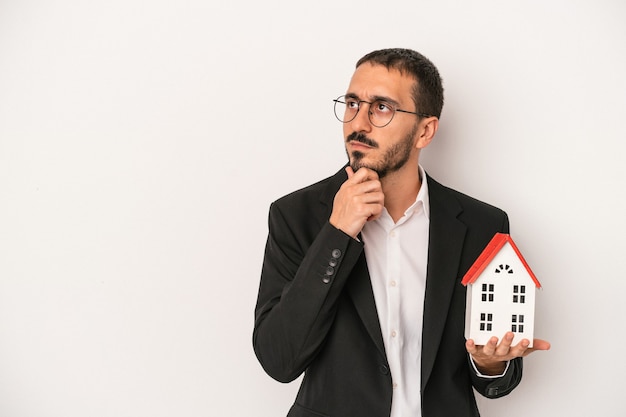 Jeune homme d'agent immobilier tenant une maison modèle isolée sur fond blanc regardant de côté avec une expression douteuse et sceptique.