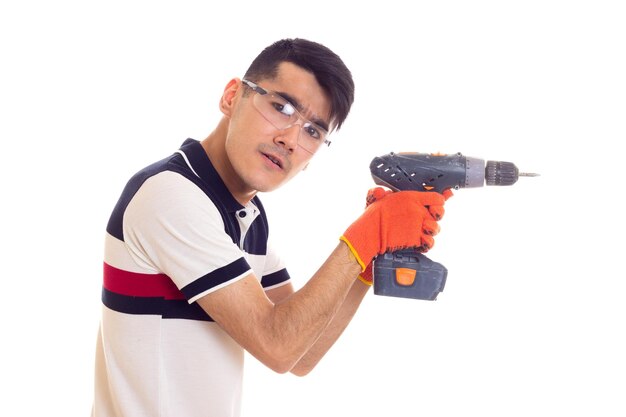 Jeune homme agréable aux cheveux noirs en t-shirt blanc et bleu avec des gants orange et des lunettes de protection tenant un tournevis électrique gris sur fond blanc en studio.