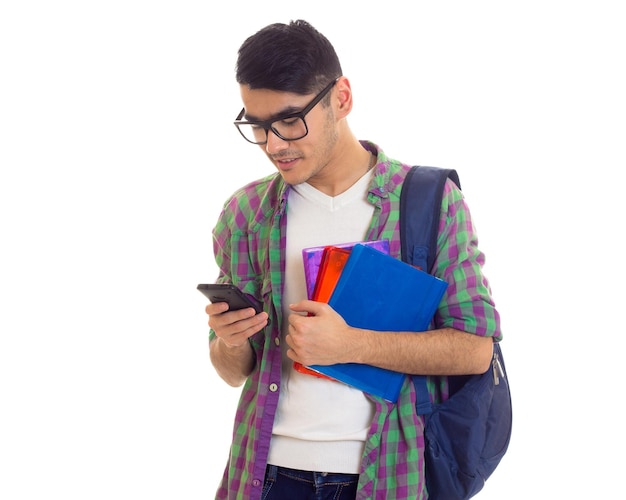 Jeune homme agréable en chemise à carreaux avec sac à dos bleu tenant des livres et utilisant un smartphone