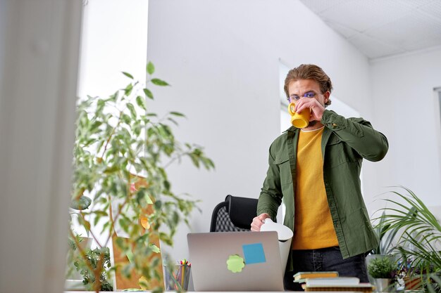 Jeune homme agréable debout derrière le bureau, boire du thé, faire une pause au travail