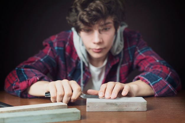 Jeune homme aiguisant un couteau sur une table en bois sur fond sombre b