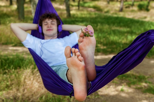 Un jeune homme allongé dans un hamac violet écoute de la musique sur des écouteurs tout en appréciant la nature