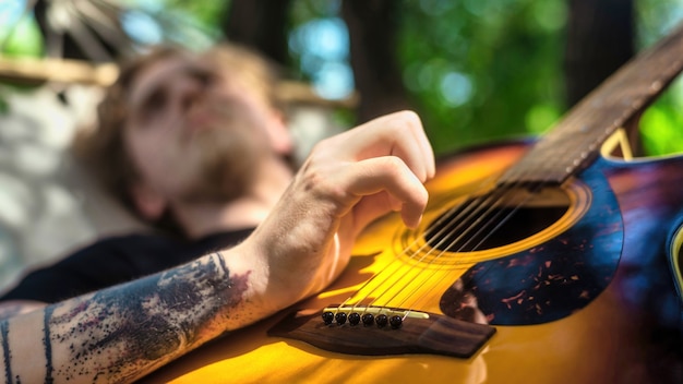 Jeune homme allongé sur un hamac joue de la guitare. Verdure autour. Glamping