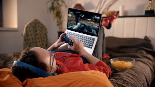 Jeune homme allongé sur le lit jouant au jeu vidéo de course sur l'ordinateur portable avec manette de jeu