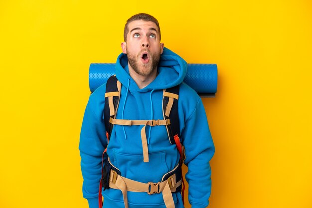 Jeune homme alpiniste caucasien avec un gros sac à dos isolé sur fond jaune à la recherche et avec une expression de surprise