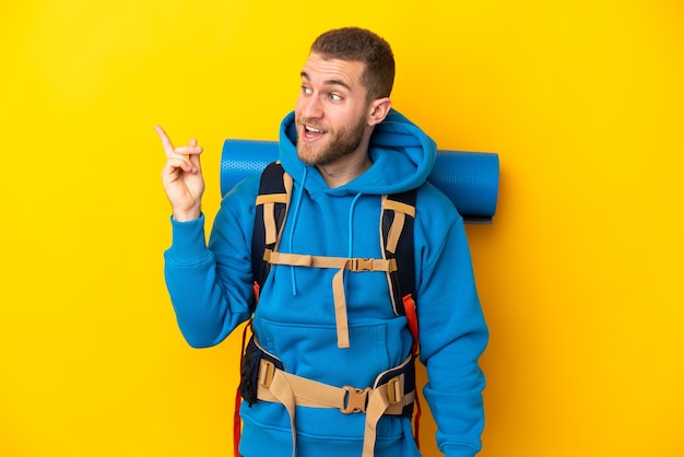 Jeune homme de l'alpiniste du Caucase avec un gros sac à dos isolé sur un mur jaune dans l'intention de réaliser la solution tout en soulevant un doigt