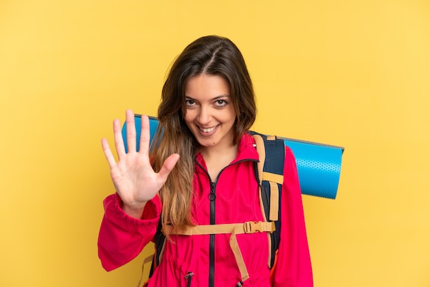 Jeune homme d'alpiniste avec un grand sac à dos d'isolement sur le fond jaune comptant cinq avec des doigts