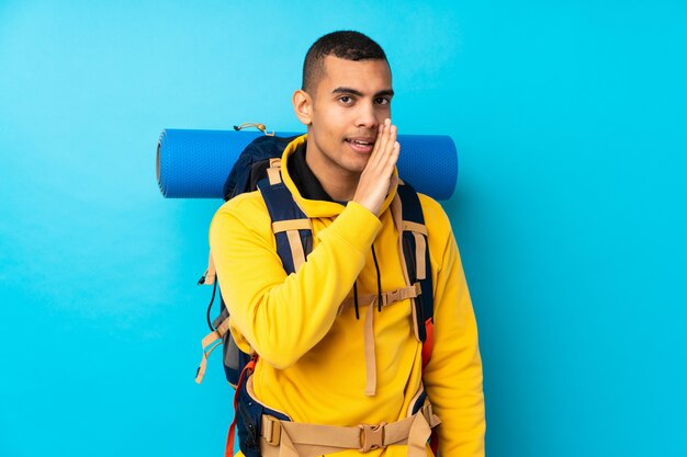 Jeune homme alpiniste avec un gros sac à dos sur mur bleu isolé chuchoter quelque chose