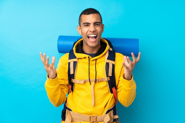 Jeune homme alpiniste avec un gros sac à dos sur le mur bleu isolé malheureux et frustré par quelque chose