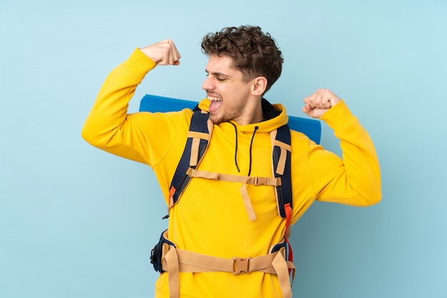 Jeune homme alpiniste avec un gros sac à dos sur le mur bleu pour célébrer une victoire