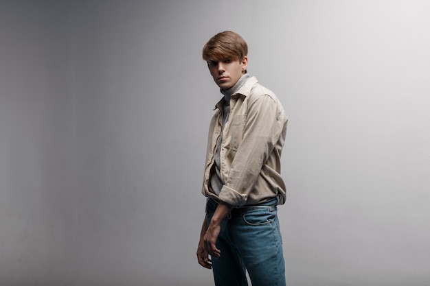 Jeune homme américain avec une coiffure à la mode dans une veste légère, légère et élégante dans un style rétro en pantalon vintage bleu se tient dans le studio près d'un mur blanc. Beau mec. Style de jeunesse moderne