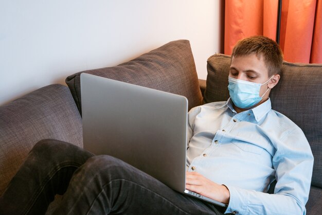 Un jeune homme d'apparence caucasienne vêtu d'une chemise bleue et d'un masque de protection avec un ordinateur portable sur le canapé...