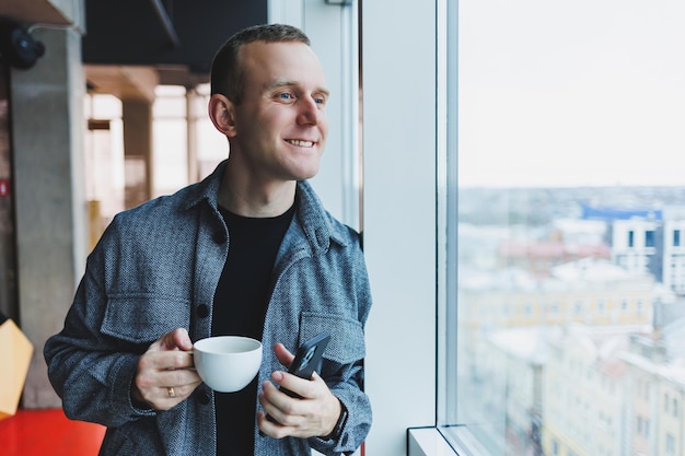 Un jeune homme d'apparence européenne en costume décontracté boit du café et regarde par la fenêtre