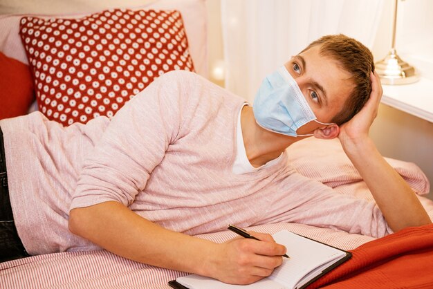 Photo un jeune homme d'apparence européenne en tenue décontractée dans un masque de protection, avec une expression pensive et rêveuse sur le visage, se trouve avec un cahier et écrit avec un stylo dedans