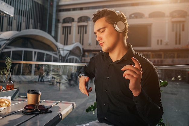 Jeune homme appréciant écouter de la musique avec des écouteurs. Il danse et agite les mains. Guy est assis dehors à table. Le soleil brille.