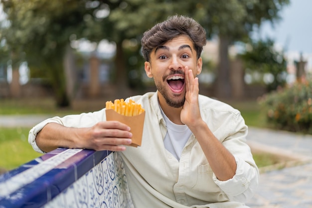 Jeune homme arabe beau tenant des chips frites à l'extérieur criant avec la bouche grande ouverte