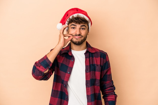 Jeune homme arabe célébrant Noël isolé sur fond beige avec les doigts sur les lèvres gardant un secret.