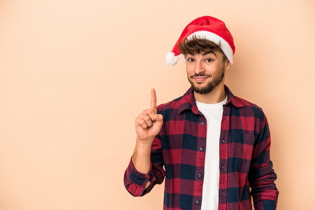 Jeune homme arabe célébrant Noël isolé sur fond beige montrant le numéro un avec le doigt.