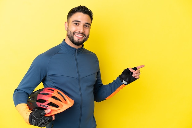 Jeune homme arabe cycliste isolé sur fond jaune, pointant le doigt sur le côté