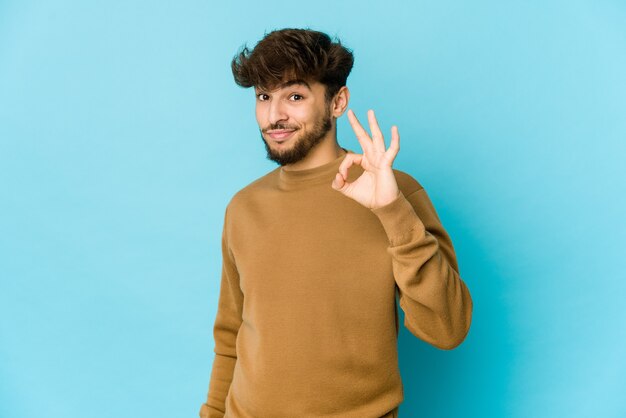 Jeune homme arabe sur fond bleu fait un clin d'œil et tient un bon geste avec la main.