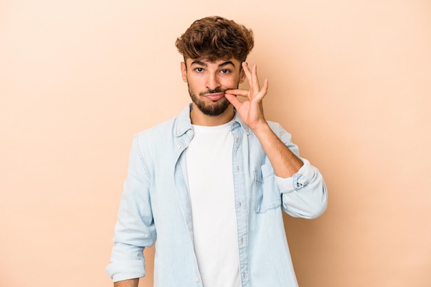 Jeune homme arabe isolé sur fond beige avec les doigts sur les lèvres gardant un secret.