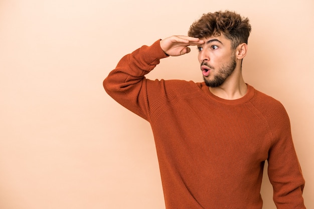 Jeune homme arabe isolé sur fond beige regardant loin en gardant la main sur le front.