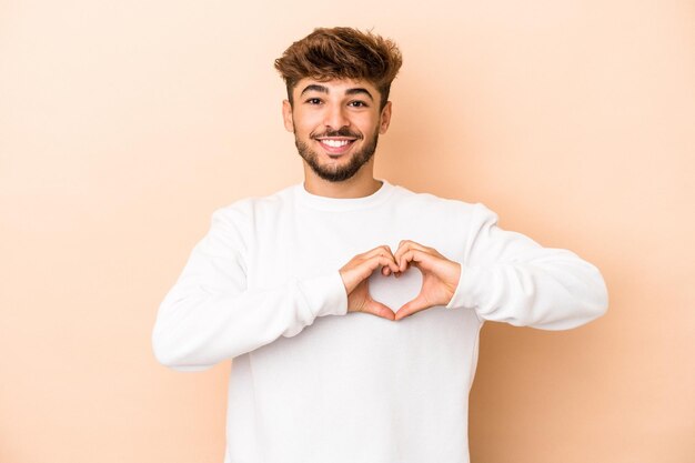 Jeune homme arabe isolé sur fond beige souriant et montrant une forme de coeur avec les mains.