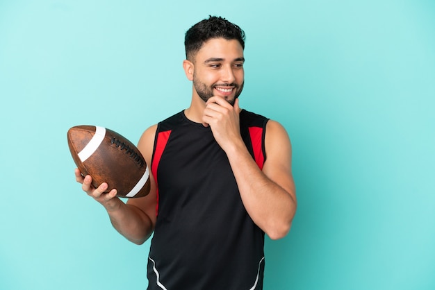 Jeune homme arabe jouant au rugby isolé sur fond bleu regardant sur le côté et souriant