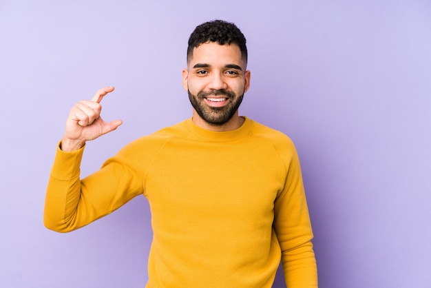 Jeune homme arabe métisse isolé tenant quelque chose de peu avec l'index, souriant et confiant.