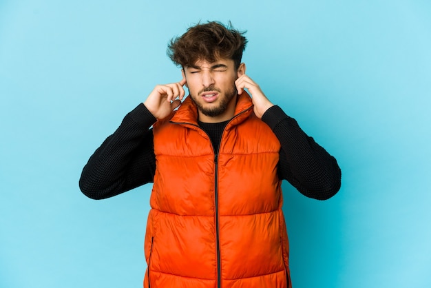 Jeune homme arabe sur un mur bleu couvrant les oreilles avec les doigts, stressé et désespéré par un bruit ambiant.