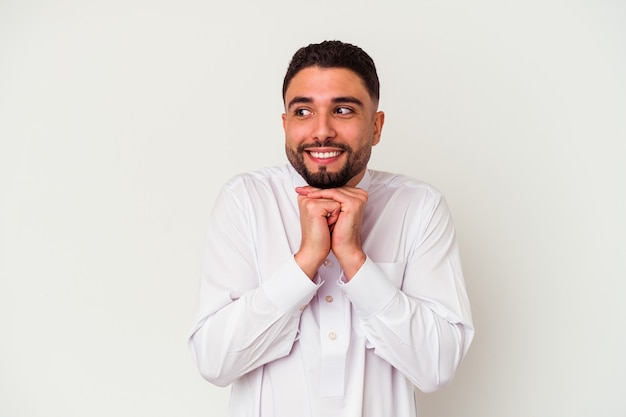 Jeune homme arabe portant des vêtements arabes typiques isolés sur blanc garde les mains sous le menton, regarde joyeusement de côté.