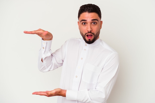 Jeune homme arabe portant des vêtements arabes typiques isolés sur fond blanc choqué et étonné tenant un espace de copie entre les mains.