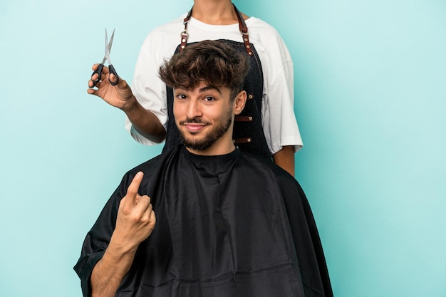 Photo jeune homme arabe prêt à se faire couper les cheveux isolé sur fond bleu pointant le doigt vers vous comme s'il vous invitait à vous rapprocher.