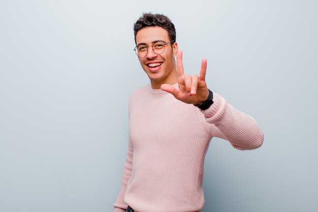 Jeune homme arabe se sentir heureux, amusant, confiant, positif et rebelle, faisant du rock ou du heavy metal signe avec la main contre le mur gris