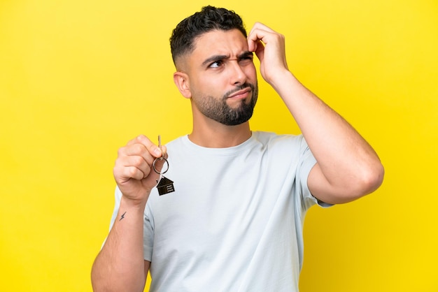 Jeune homme arabe tenant les clés de la maison isolé sur fond jaune ayant des doutes et avec une expression de visage confuse