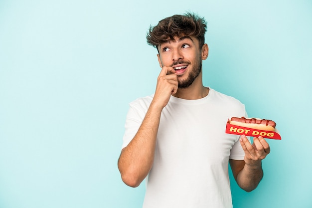 Jeune homme arabe tenant un hot-dog isolé sur fond bleu détendu en pensant à quelque chose en regardant un espace de copie.