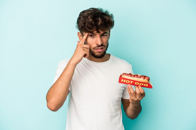 Jeune homme arabe tenant un hot-dog isolé sur fond bleu montrant un geste de déception avec l'index.