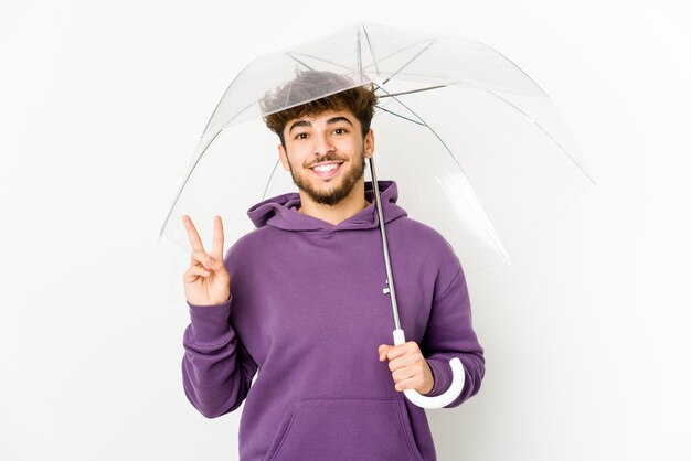 Jeune homme arabe tenant un parapluie joyeux et insouciant montrant un symbole de paix avec les doigts