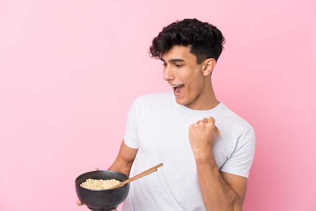 Jeune homme argentin sur mur blanc isolé célébrant une victoire tout en tenant un bol de nouilles avec des baguettes