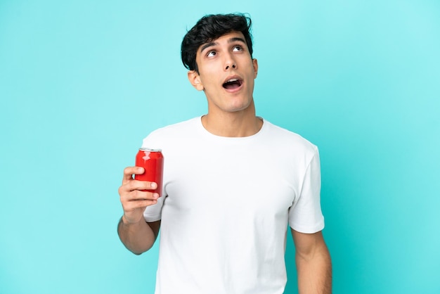 Jeune homme argentin tenant un rafraîchissement isolé sur fond bleu en levant et avec une expression surprise