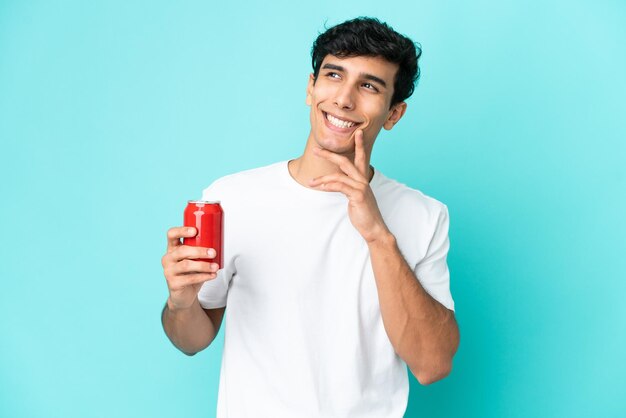 Jeune homme argentin tenant un rafraîchissement isolé sur fond bleu en levant tout en souriant