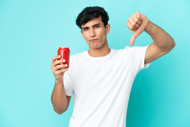 Jeune homme argentin tenant un rafraîchissement isolé sur fond bleu montrant le pouce vers le bas avec une expression négative