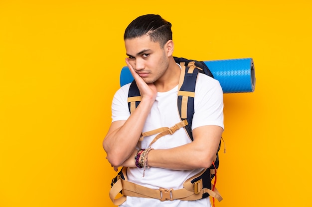 Jeune homme asiatique alpiniste avec un gros sac à dos isolé sur fond jaune malheureux et frustré