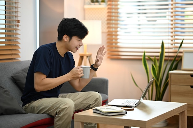 Jeune homme asiatique assis sur un canapé et faisant un appel vidéo ayant une conversation virtuelle sur un ordinateur portable depuis la maison