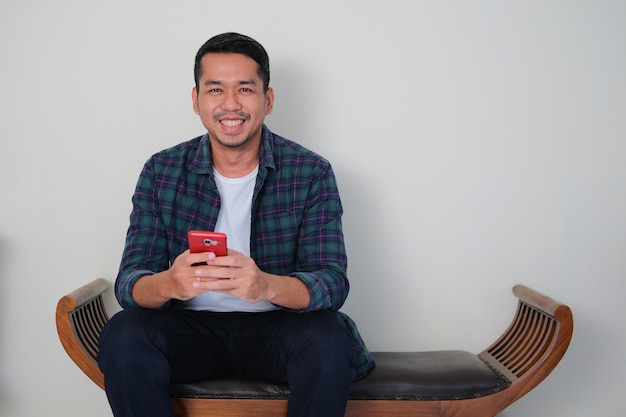 Jeune homme asiatique assis sur une chaise en bois tenant un téléphone portable avec une expression de visage heureux