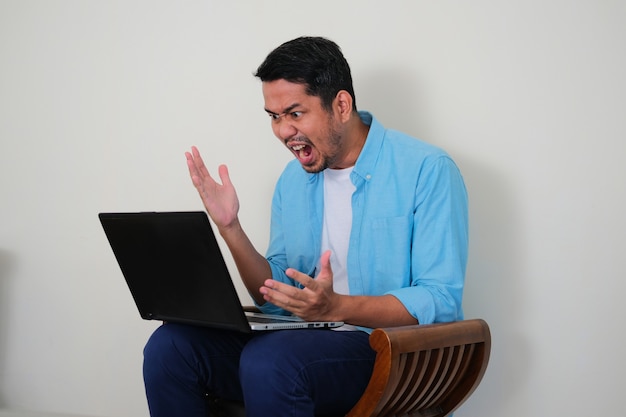 Jeune homme asiatique assis sur une chaise et regardant son ordinateur portable avec une expression de rage