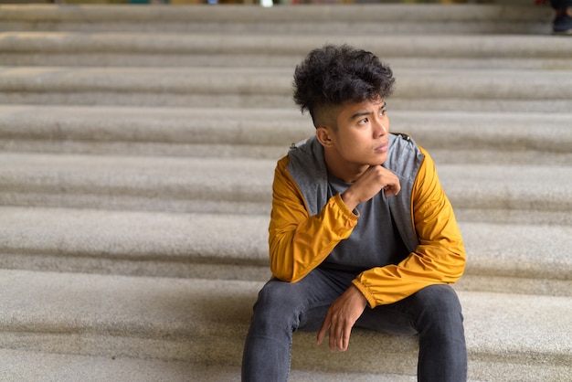 Jeune homme asiatique aux cheveux bouclés pensant et assis sur l'escalier dans la ville