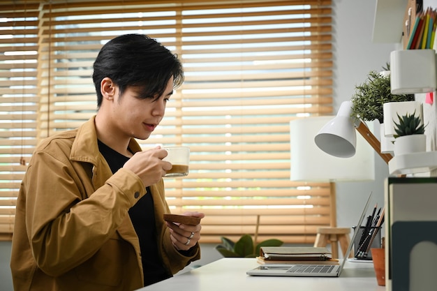 Jeune homme asiatique buvant du café et lisant des e-mails sur un ordinateur portable.