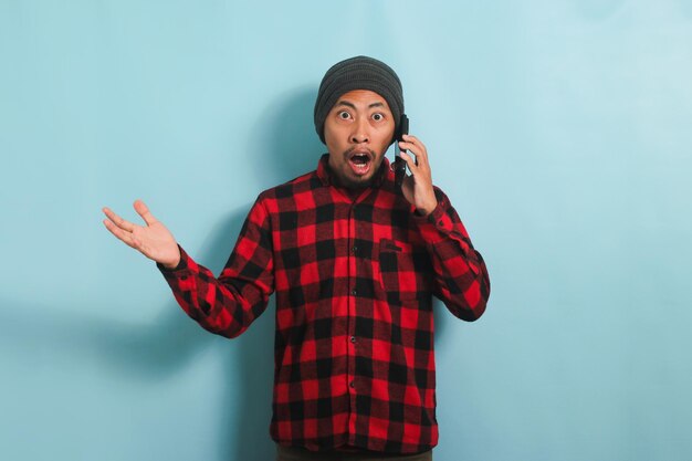 Un jeune homme asiatique choqué est surpris en parlant sur son téléphone portable isolé sur fond bleu