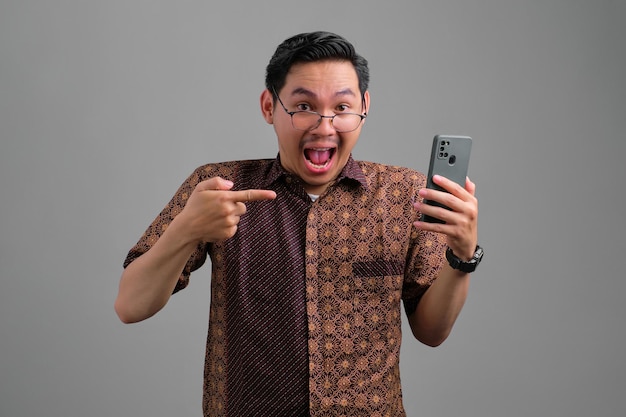 Jeune homme asiatique choqué portant une chemise batik pointant du doigt un smartphone isolé sur fond gris